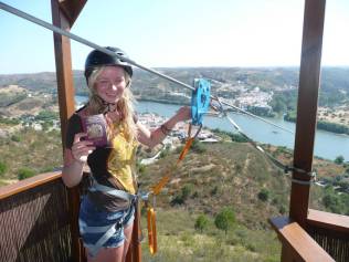 PICS BY LIMITZERO / CATERS NEWS - (PICTURED: A British tourist poses at the top of the hipline between Spain and Portugal with her passport) - A ZANY new zipline whisks travellers from Spain to Portugal in less than one minute. The border-crossing attraction is the first - and currently only - zipline in the world to transport users across a border. Thrillseekers board in Sanlucar de Guadiana in Andalucia, Spain, before whizzing down the line at around 50mph - and landing across the Guadiana River in Alcoutim, Portugal. And thanks to time zone differences between the two nations, the 60 second journey will see the zipliners travel back in time one hour. SEE CATERS COPY.
